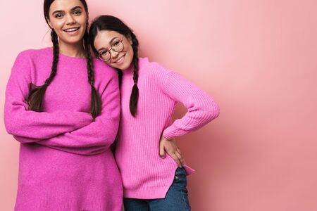Image of two happy teenage girls with braids in casual clothes smiling at camera isolated over pink...