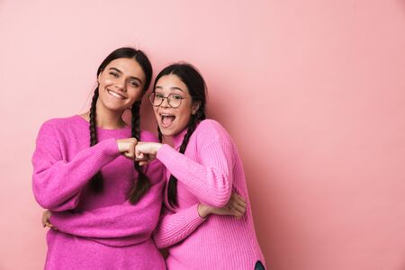 Image of two young teenage girls with braids smiling and bumping their fists together isolated over pink backgroundの写真素材