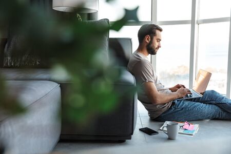 Image of a concentrated young man indoors at home using laptop computer sit on floor.の写真素材