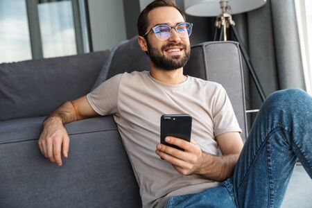 Attractive smart young man sitting on a floor in the living room, using mobile phone
