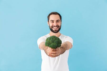 Portrait of a young cheerful excited bearded man wearing t-shirt standing isolated over blue background, showing broccoliの写真素材