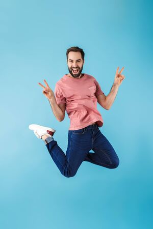 Full length of a young cheerful excited bearded man wearing t-shirt jumping isolated over blue...