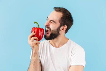 Handsome cheerful young bearded man wearing t-shirt standing isolated over blue background, biting...