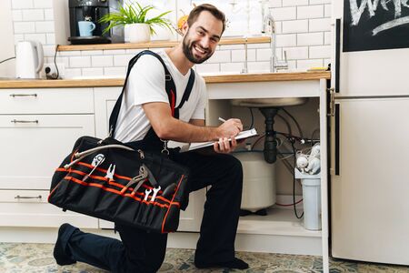 Image of young caucasian plumber man in uniform holding bag with equipment and clipboard while...