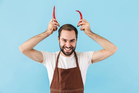 Angry young bearded man cook wearing apron standing isolated over blue background, holding chilli pepper at his head pretending devil hornsの写真素材