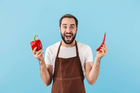 Cheerful young bearded man wearing apron standing isolated over blue background, holding red...
