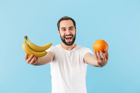 Handsome cheerful young bearded man wearing t-shirt standing isolated over blue background, showing orange and bananaの写真素材