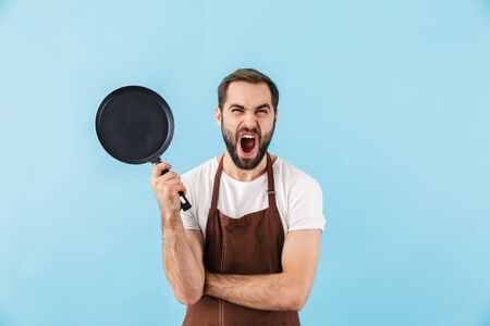 Photo of young screaming displeased man chef isolated over blue wall background holding frying pan.の写真素材