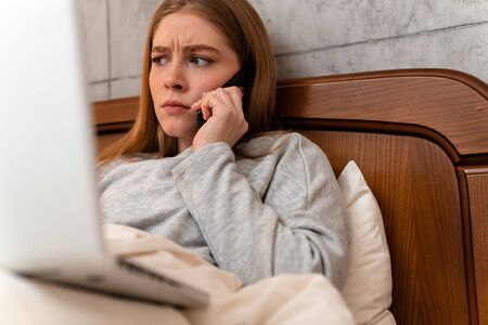 Image of a shocked displeased young woman indoors at home in bed using laptop computer talking by mobile phone.の写真素材