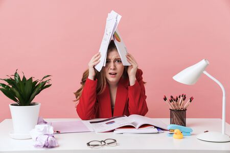Image of a confused negative young woman sit at the table indoors isolated over pink wall background holding documents.の写真素材