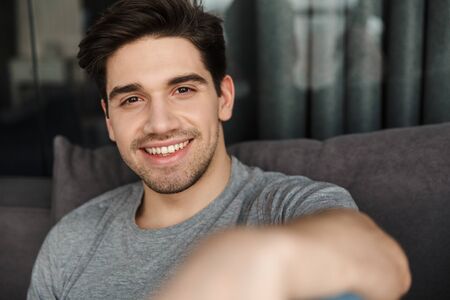 Portrait of an attractive smiling young bearded man wearing casual clothes sitting on a couch at the...