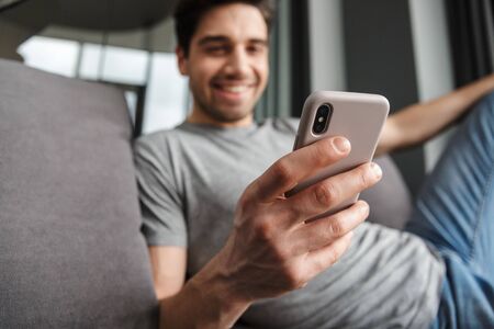 Portrait of an attractive smiling young bearded man wearing casual clothes sitting on a couch at the living room, using mobile phoneの写真素材