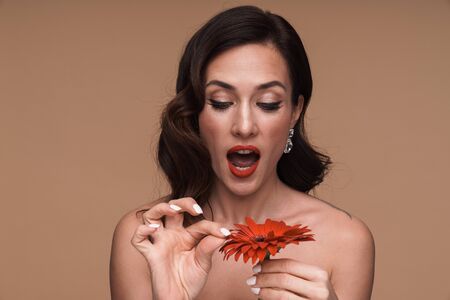 Beauty portrait of brunette adult half-naked woman holding red flower and pulling out petals isolated over beige backgroundの写真素材