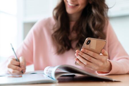 Cropped photo of a cheerful beautiful young woman sit indoors at home using mobile phone writing notes.の写真素材