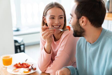 Portrait of young happy couple eating together at table while having breakfast in cozy kitchen at homeの写真素材