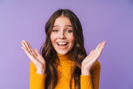 Image of young beautiful woman with long brown hair smiling and raising hands in delight isolated over violet backgroundの写真素材