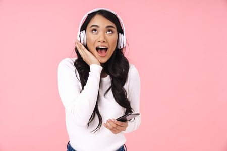 Cheerful pretty young asian woman listening to music with wireless headphones isolated over pink background, holding mobile phoneの写真素材