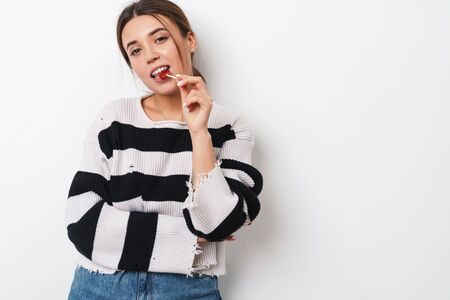 Image of young pleased woman in striped sweater licking lollipop and looking at camera isolated over...