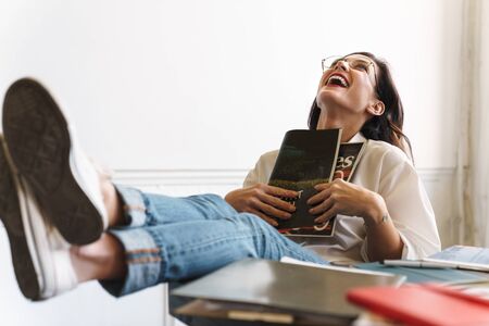 Lovely young brunette woman reading magazine while relaxing at the table in the cafe indoors, laughingの写真素材
