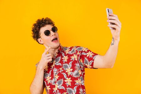 Image of young delighted man taking selfie and pointing finger at cellphone isolated over yellow backgroundの写真素材
