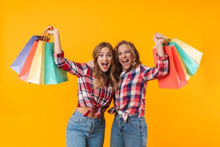 Image of two young beautiful girls wearing plaid shirts smiling and holding colorful shopping bags isolated over yellow backgroundの写真素材