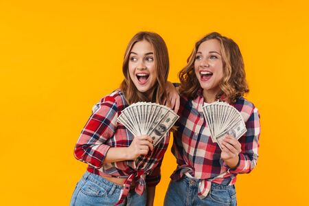 Image of two young beautiful girls wearing plaid shirts holding dollar banknotes isolated over yellow backgroundの写真素材