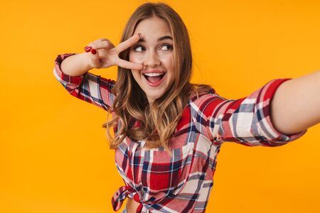 Image of happy young attractive woman wearing plaid shirt smiling while taking selfie photo isolated over yellow backgroundの写真素材