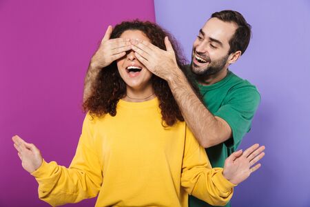 Photo of happy man in colorful clothing smiling and covering woman's eyes standing behind her isolated over violet backgroundの写真素材