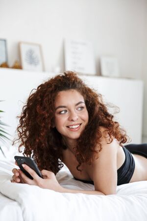 Image of a cheerful happy young lady in lingerie indoors in home hotel on bed using mobile phone.の写真素材