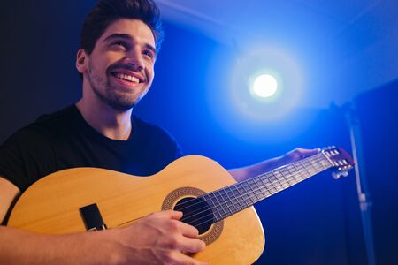 Image of a handsome happy young cheery man musician play on a guitar on scene in night club over dark background with flash lights.の写真素材