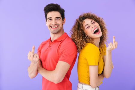 Portrait of cheerful caucasian people man and woman in basic clothing smiling and showing middle fingers at camera isolated over violet backgroundの写真素材