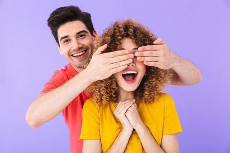 Photo of cheerful couple, man smiling and covering woman's eyes standing behind her isolated over...