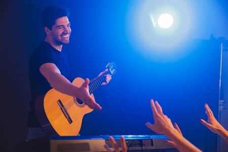 Image of a handsome happy young positive man musician play on a guitar on scene in night club over dark background with flash lights give a hand to fans.の写真素材