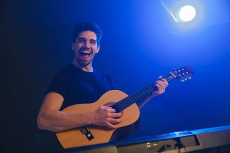Image of a handsome happy young positive man musician play on a guitar on scene in night club over dark background with flash lights.の写真素材