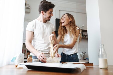 Image of joyous couple rejoicing while cooking breakfast and mixing dough in kitchen at homeの写真素材