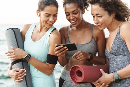 Three cheerful young sport girls standing at the beach, holding fitness mats, using mobile phoneの写真素材