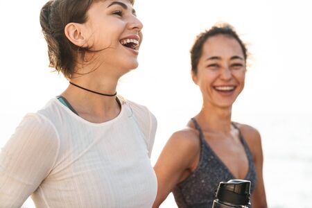 Image of joyful fitness women in sportive clothes smiling and standing by seaside in morning