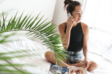 Portrait of gorgeous woman in summer clothes talking on mobile phone while reading magazine on bed...