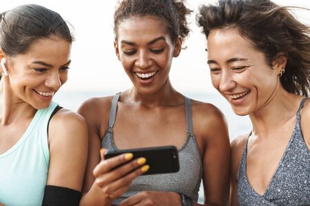 Close up of three cheerful young sport girls standing at the beach, holding fitness mats, using mobile phoneの写真素材