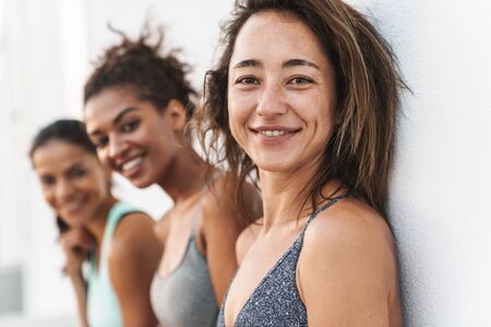 Photo closeup of healthy multiethnic sportswomen in tracksuits smiling at camera and standing over white wall outdoorsの写真素材