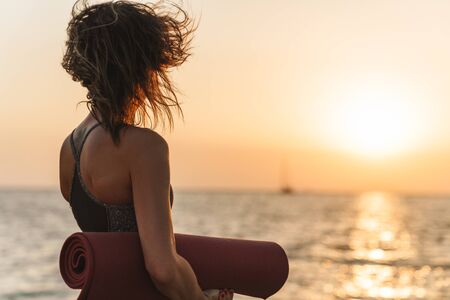 Photo from back of caucasian sportswoman in tracksuit looking away while standing with yoga mat by...