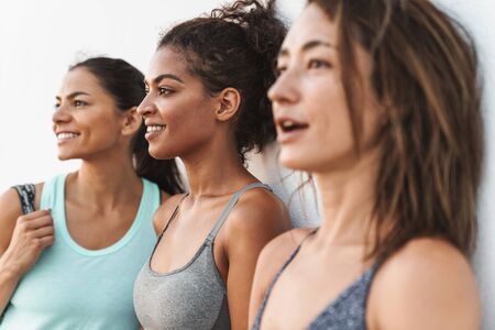 Photo closeup of joyful multiethnic sportswomen in tracksuits smiling at camera and standing over white wall outdoorsの写真素材