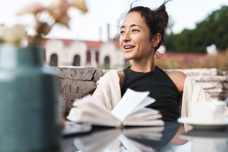 Portrait of pleased brunette woman in casual clothes reading book and drinking coffee while resting...