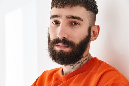 Image closeup of young bearded man with nose jewelry wearing orange shirt standing over white wall...