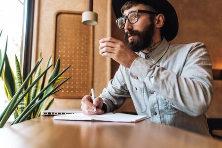 Photo of serious young man wearing eyeglasses making notes in diary while drinking coffee in cafe indoorsの写真素材