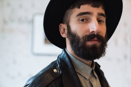 Photo closeup of bearded young man wearing hat posing and looking at camera in bright room indoorsの写真素材
