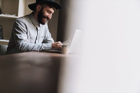 Photo of laughing young man wearing hat typing on laptop and drinking coffee while sitting at table...