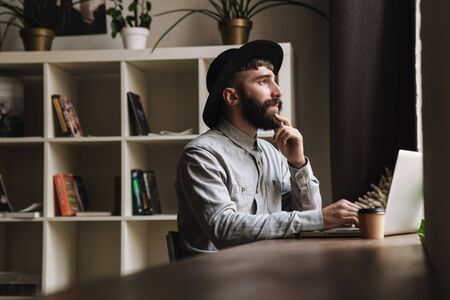Photo of thinking young man wearing hat typing on laptop and drinking coffee while sitting at table in cafe indoorsの写真素材