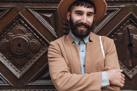 Attractive smiling young man wearing autumn coat and hat standing outdoors at the city street in front of the wooden doorの写真素材