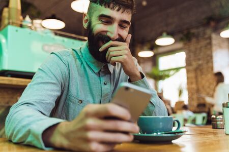 Attractive smiling young bearded man wearing shirt using mobile phone while sitting at the cafe indoors and drinking coffeeの写真素材